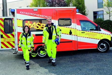 Eine Frau und ein Mann im gelben Schutzanzuf vor einem Fahrzeug des ASB.